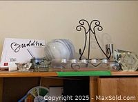 Photo of the lot showing Buddha Board box, large round glass bowl, assorted glass candle holders, black ornate metal picture holder, and hexagonal glass accent piece all arranged on shelf.