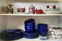 Photo showing blue dinner plates, aide plates, and blue bowls stacked inside cabinet. Upper shelf shows assorted porcelain bowls, jars, and teacup.