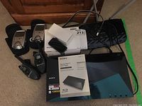 Photo showing the Sony Blu-ray DVD player with manual, two Motorola walkie talkies, Omichron hearing assist device, and the TP-Link router all laid on carpet.