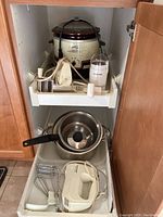 Kitchen cabinet drawers containing the Crock-Pot slow cooker on top pull-out shelf, electric coffee grinder, handheld cheese grater, and lower drawer with stainless steel pots and Black & Decker electric hand mixer with manual.