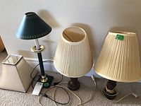 Two wooden table lamps with one green metal lampshade and two spare fabric shades on floor in front of wall.