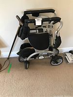 Side view of silver Evolution Xpresso Lite Mini C walker folded, showing black cushioned seat and backrest, four wheels, padded handles with brake controls, attached storage bag, with wooden cane resting on it.