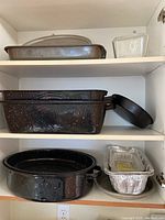 Kitchen cabinet shelf showing a rectangular steel roasting pan, clear glass loaf pan, two foil pans, one oval dark speckled enamel roasting pan with lid, and a round pizza pan beneath, arranged on white shelves.