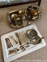 Wide shot of the silver plate tray holding two wine goblets and tea service including teapot, sugar bowl, and creamer, with small pewter leaf-shaped dish and spoon nearby, and various cutlery laid out on a white tray.