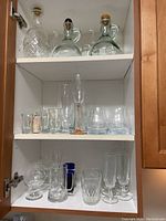 Top cabinet shelves with assorted drinking glasses including a champagne flute, tumblers, stemmed glasses, blue handled mug, and three decorative oil bottles with stoppers.