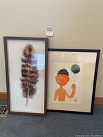Two framed artworks leaning against a wall, one is a watercolor painting of a person holding a balloon by Mabee and the other is a large feather painted on glass in a shadow frame.