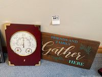 Two items on floor against wall: a rectangular wood framed barometer with brass corner accents and a wooden sign with painted text