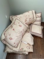 Photo showing the comforter, pillows, bed skirt, and throw pillows neatly stacked on floor in neutral lighting.
