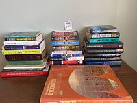 Photo shows a stacked assortment of books part of the lot including the World Atlas, novels by John Grisham, Lee Child, James Patterson, biography and memoir titles, and sports-themed books.