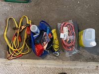 Photo showing yellow jumper cables, blue mesh bag with small items, thick yellow towing rope with hooks, coiled red jumper cables in plastic, a folded black blanket, and yellow fluid container.