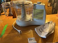 Hamilton Beach food processor with clear plastic bowl, metal blade insert, and white motor base with control dial; includes instruction manual folded beside the appliance.
