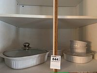 White rectangular rimmed casserole with glass lid and two ramekins stacked on top of an additional round casserole without a lid on shelf