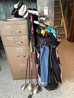 Golf bag standing on floor with clubs inside and some clubs propped outside. Bag is black with pink and several compartments, includes headcovers in white and black.