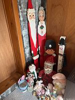Wide view of wooden Santa, Mrs. Claus, nutcracker figure, wire red Santa, and pastel Easter decorations on floor against wooden walls.
