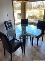 Round heavy glass kitchen table with four black vinyl Parson chairs around it, shown in a room with tiled floor and window light.