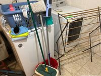 Photo showing laundry basket filled with cleaning cloths and bottles, mop and bucket next to it, broom with mop head, drying rack, and shoe shining items on top of washing machine.