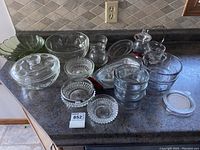 Photo showing collection of various clear and green Anchor Hocking glass bowls, candy dishes, serving bowls including items with lids, rose-shaped bowl, creamer, and sugar bowl.