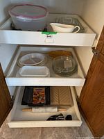 View of cabinet shelves holding a variety of kitchen items including glass plates, plastic storage container, white ceramic dishes, yellow dish, marble rolling pin, metal BBQ basket, metal trays and various kitchen utensils.
