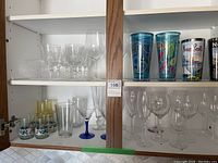 Cabinet shelf with various glasses: six pinwheel wine glasses, other clear wine glasses, short glasses with prints, and colorful plastic tumblers