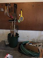Overview of gardening tools in a green garbage can placed beneath a pegboard wall. Tools include shovels, broom, and braces. Coiled garden hose and metal live trap nearby on floor.