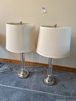 Pair of table lamps with fabric shades and chrome bases, standing on carpet in front of a wall.
