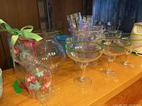 Photo showing various plastic glasses including margarita-style glasses with green rims and clear stems and bases, a clear plastic ice bucket, and other assorted drinkware on a wooden surface.