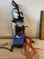 Shows full view of blue Simoniz pressure washer with coiled black hose and orange extension cord in front.