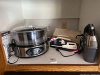 Shows all four kitchen appliances together on a shelf: Black & Decker steamer/rice cooker, Hamilton Beach hand mixer in box, Moulinex cutter, and a small handheld appliance