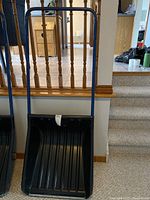 Full view of the snow pusher showing the black plastic wide blade and blue metal handle in the indoor setting.