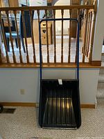 Front view of a heavy duty plastic snow remover with a black scoop and a blue metal handle with a horizontal grip bar, standing upright on a carpeted floor with wooden stair railing in the background.