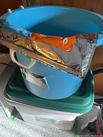 Blue large utility bucket with white handles sitting on top of two white plastic storage totes with green and dark green lids. Thermal bag is resting folded on bucket.