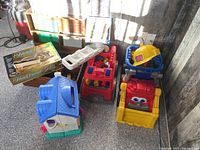Photo of handcrafted wooden box containing assorted LEGO-type bricks and green and white base plates, and toy vehicles including a red fire truck and yellow dump truck, along with doll house and boxed puzzle.