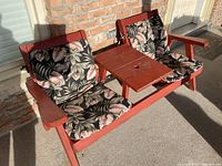 Wood bench showing two seat cushions with floral pattern, center table with umbrella hole, reddish wood paint, worn condition