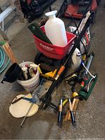 General overview of the entire lot showing EarthWay spreader, white sprayer bottle, bucket with tools, hedge cutter, Fiskars weeder, and other gardening tools and accessories.