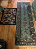 Two area rugs side by side on wood floor. Left rug is multicolor hooked wool rug with black border; right rug is teal colored oriental style runner with floral and geometric motifs and fringe on short edges.