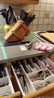 Kitchen countertop with wooden knife block holding knives and honing rod, container with black spatulas and ladles, folded cloth, and open drawers containing organized silverware including spoons and forks.