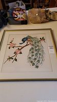 Framed needlepoint artwork of a peacock on a branch with pink flowers, full view showing frame and surrounding items.