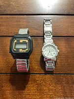 Two watches on wooden surface: one Bulova silver-tone chronograph with white face and crystals, one vintage digital watch with black face and metal band