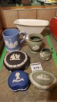 Photo showing full lot arrangement on a surface including the various Wedgwood jasperware cups, vases, and trinket boxes along with a white ribbed container in the back.