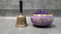 Photo of the vintage brass bell standing next to the purple Tibetan singing bowl with gold inscriptions and a purple felt striker inside.
