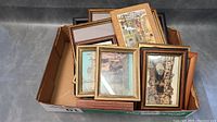Box containing multiple small framed paintings and prints with visible frames of wood, gold, and corkboard showing urban scenes and landscapes.