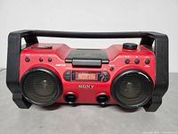 Front view of red Sony Carpenter boombox showing large dual speakers, digital display tuned to FM 107.9, control buttons and dial, with black handle on top.