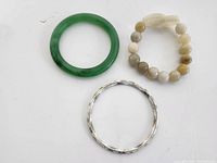 Top-down image of three bangles on white background. Green glass bangle, beaded stretch bangle with carved cream and brown beads, and silver-tone metal enamel bangle shown clearly.