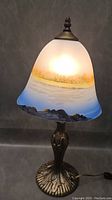 Front and side view of the vintage table lamp illuminated, showing painted landscape glass shade and decorative metal base.