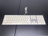 Apple wired keyboard with numeric keypad, white keys, aluminum body, USB cable coiled on top, placed on black surface.