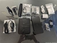 Top view of assorted phone and car chargers, phone clip holder, two flashlights, leather phone pouch and accessories arranged on glass surface.