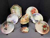 Set of six hand-painted dishes arranged with four floral plates forming a semi-circle and butter dish and handled tray in front, showing variety of floral designs, gold rims, and pastel colors.