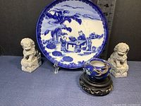 Front view of blue and white ceramic plate, pair of carved stone foo dogs, small ceramic bowl with lizard on wooden stand, ruler for scale.