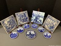 Front view showing 11 blue and white Delft-style decorative tiles and plates, various sizes and round and square shapes, depicting Dutch life scenes with windmills and figures