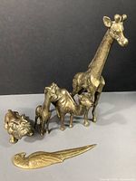 Photo showing a set of five brass items: a bulldog figurine, baby giraffe figurine, camel figurine, adult giraffe figurine, and a parrot letter opener laid out on gray surface with black backdrop.